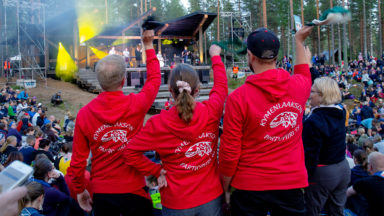 Kolme partiolaista heiluttaa huiveja Kymenlaakson Partiopiiri-hupparit päällä bändin soittaessa lavalla Johtajatulilla.