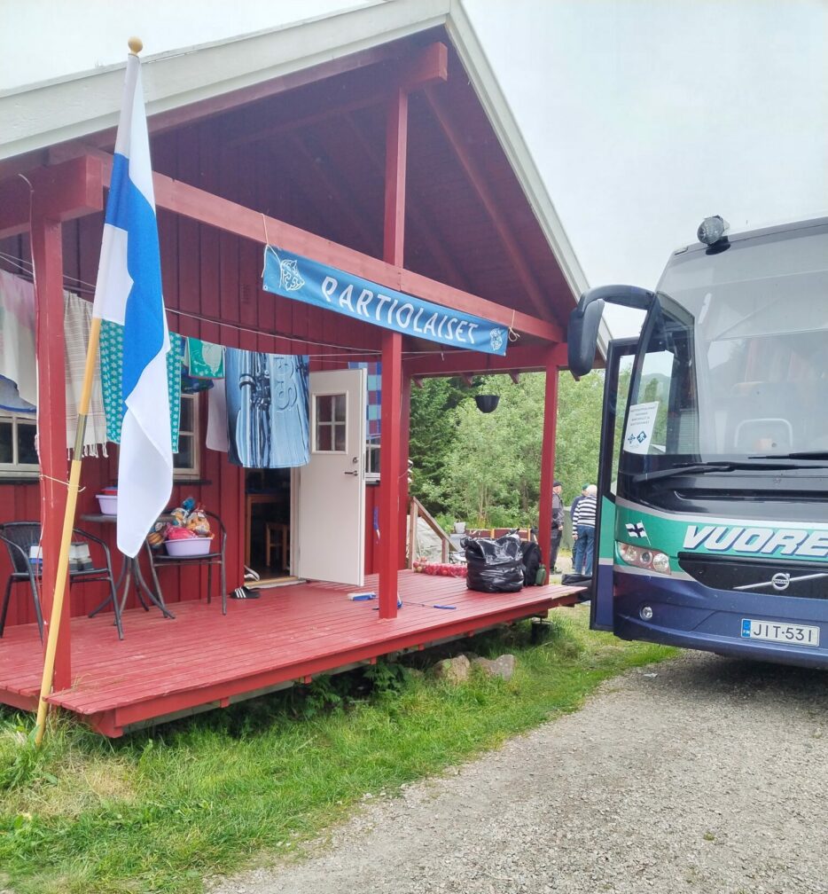 Bussi seisoo talon vieressä, jonka terassille on laitettu Suomen lippu ja partiolaisten banneri.