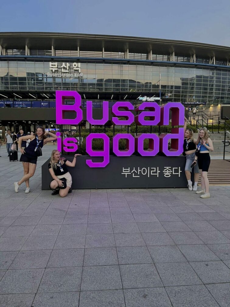 Kuvassa partiolaisia seisoo kyltin edessä, jossa lukee "Busan is good" englanniksi ja koreaksi.