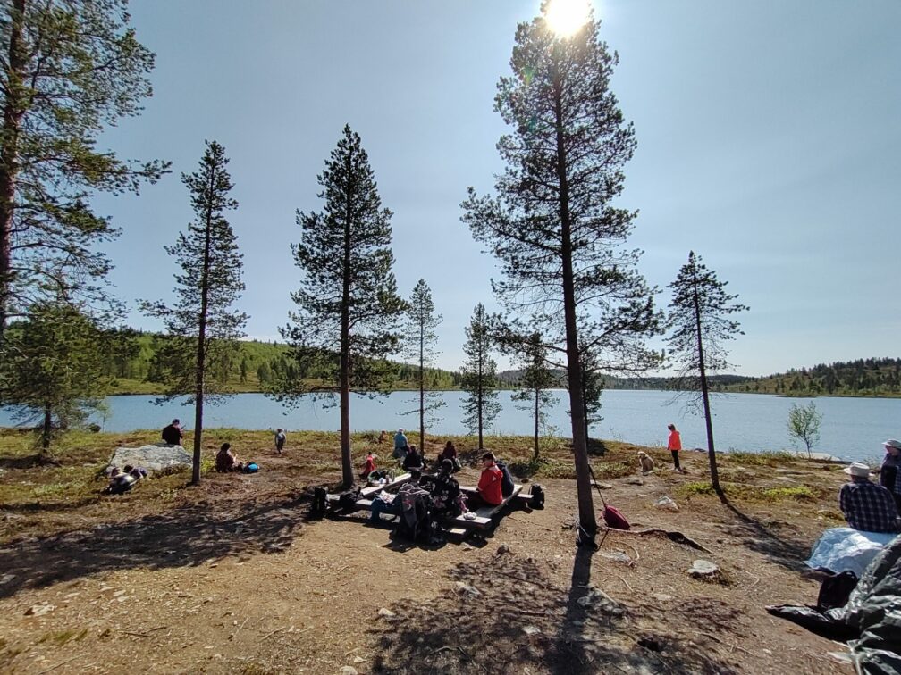 Järven rannalla nuotiopaikalla istuu useita ihmisiä.